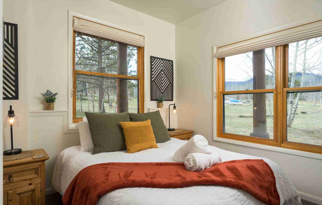 Queen bedroom with white walls, a rust-colored throw, and windows to the pines